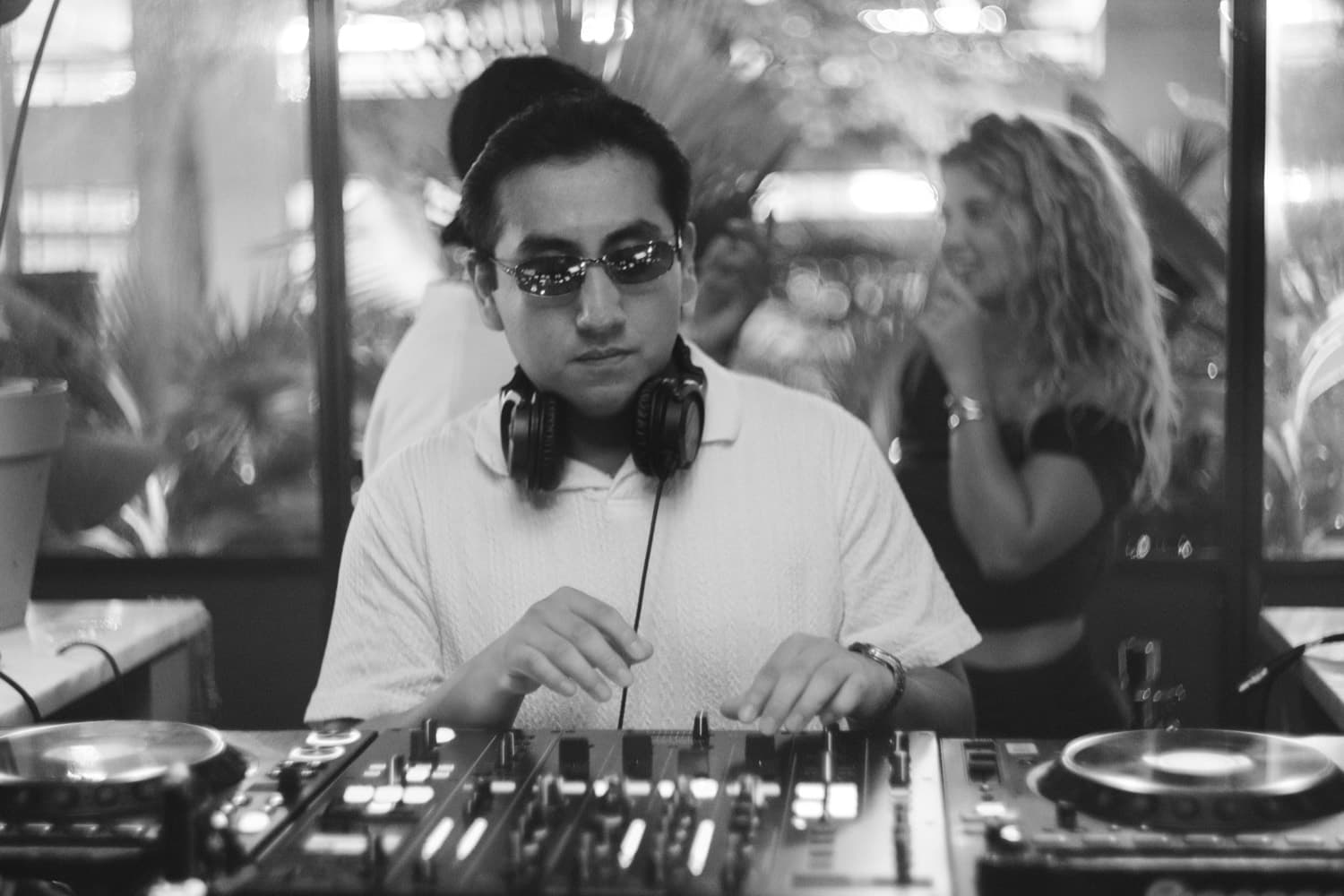 LP behind the decks in sunglasses and headphones around his neck, plants behind him