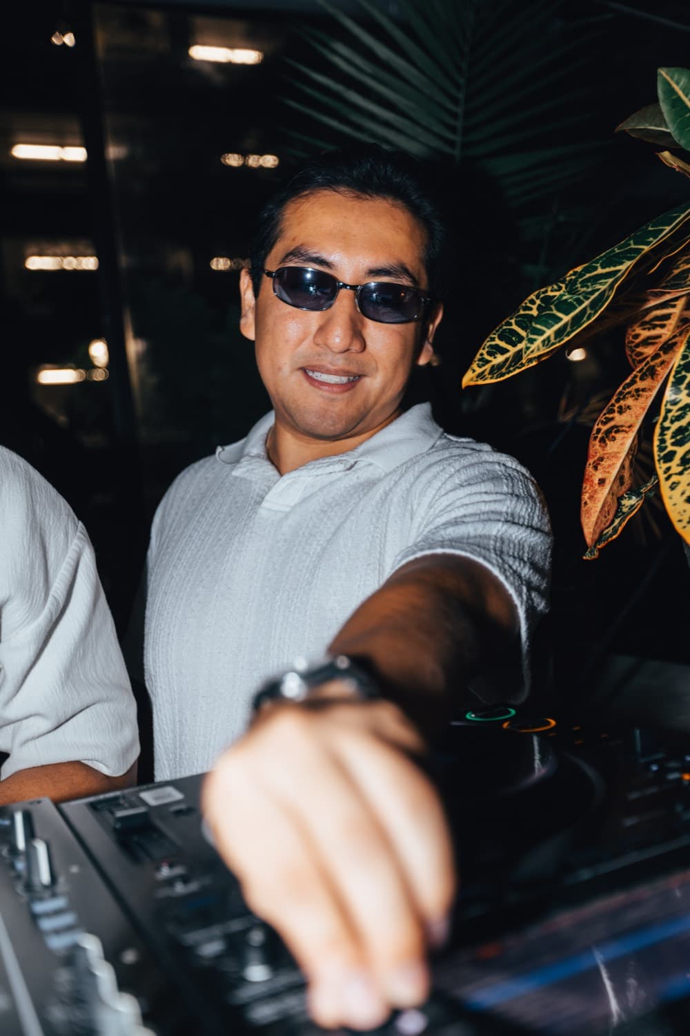 LP smiling and reaching toward the camera from behind the decks