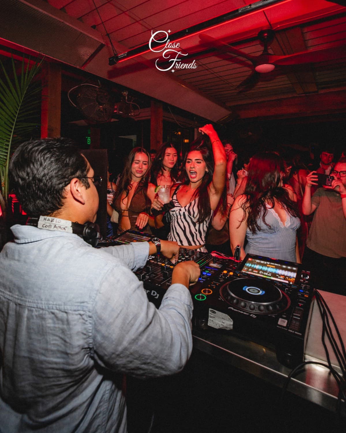 A crowd dancing with arms raised under red light near the DJ booth