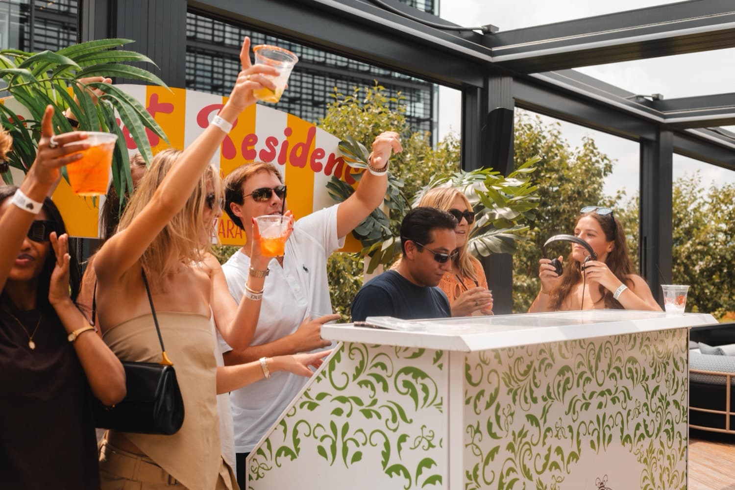 A group with arms raised holding drinks under the Velvet Residence sign at a daytime rooftop party